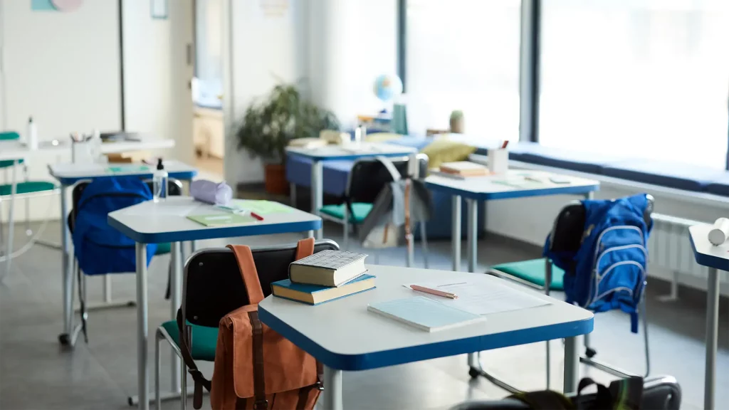 Aula con mesas escolares y mochilas colgadas en las sillas.